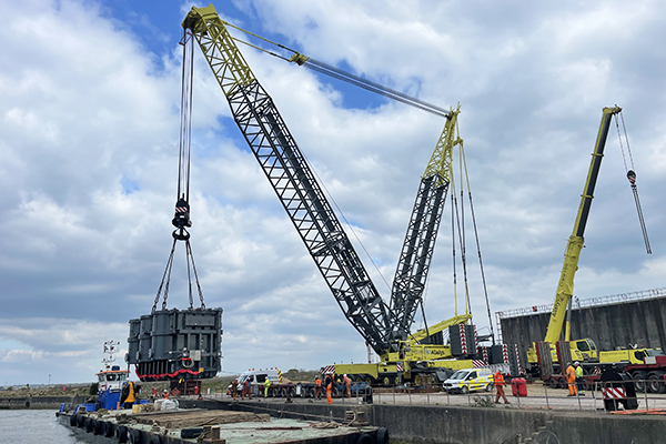 One heavy lift crane lifting a grey piece of heavy equipment from a dock